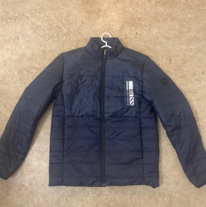 Mörkblå pufferjacka från Hugo Boss - Snygg mörkblå pufferjacka från Hugo Boss med vit logga på bröstet. Jackan har ståkrage, hel dragkedja och quiltad design. Tillverkad i polyester och har långa ärmar. Perfekt för dig som vill ha en stilren och modern look.