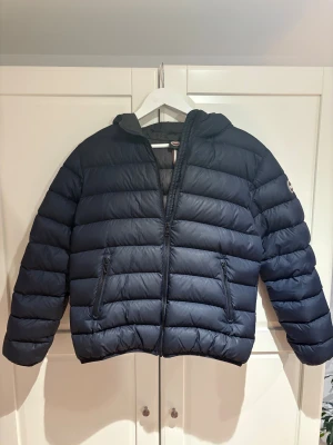Mörkblå Colmar pufferjacka med huva - Säljer en mörkblå pufferjacka från Colmar med huva och två sidofickor med dragkedja. Jackan har ett stilrent, quiltat mönster och logotyp på ärmen. Perfekt för kalla dagar. Storlek 160 vilket motsvarar S. Ny pris 5000kr mitt pris 2199!