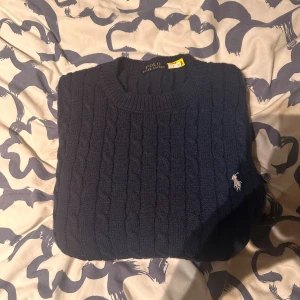 Mörkblå kabelstickad tröja Polo Ralph Lauren - Kabelstickad mörkblå tröja från Polo Ralph Lauren med klassisk rund halsringning och broderad logga på bröstet. Tröjan är i mjuk ull och har en tidlös look som passar perfekt till jeans eller chinos.