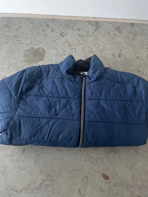 Blå pufferjacka från H&M i storlek 146 - Blå pufferjacka från H&M med ståkrage och svart innerfoder. Jackan har en rak passform, synlig dragkedja framtill och ett litet tryck med koordinater på nedre delen. Perfekt för kalla dagar och enkel att matcha med din stil. Storlek 146