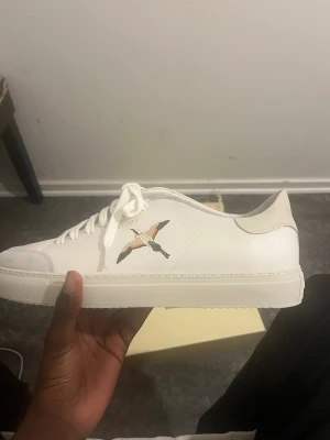 Vita Axel Arigato sneakers med broderad fågel - Snygga vita sneakers från Axel Arigato i skinn med broderad fågel på sidan. Klassisk låg modell med rund tå, platt sula och vita snören. Perfekta för dig som gillar stilrena och trendiga skor med en unik detalj.