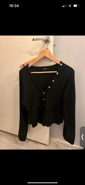 Svart kofta med knappar från Gina Tricot - Svart kofta från Gina Tricot i storlek M. Modellen har v-ringning och cool knappdetalj längs ena axeln och framtill. Långärmad och i mjukt stickat material, perfekt för lager-på-lager och snygg till både jeans och kjol.