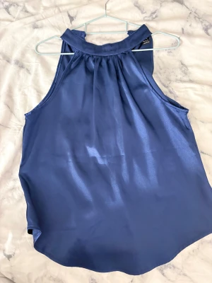 Mörkblå satinlinnetopp från SHEIN - Snygg mörkblå linne-topp i satin från SHEIN. Toppen har hög hals med två knappar i nacken och är ärmlös med lätt rynkade detaljer framtill. Perfekt för dig som gillar en stilren och elegant look.