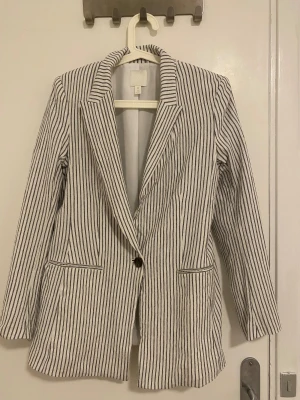 Randig kavaj från H&M, storlek 38 - Snygg vit kavaj med svarta vertikala ränder från H&M. Klassisk modell med krage, en knapp framtill och två fickor. Perfekt för dig som gillar stilrena och trendiga plagg. Materialet är mjukt och bekvämt, passar till många olika looks.