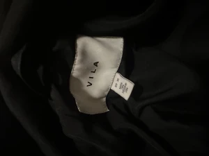 Svart lång pufferjacka från Vila - Säljer en svart lång pufferjacka från Vila med hög krage och bälte i midjan. Jackan har knappar framtill, stora fickor och en riktigt oversized look. Perfekt för kalla dagar när du vill vara både varm och snygg.