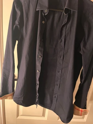 Marinblå skjorta Burberry Brit XS - Snygg marinblå skjorta från Burberry Brit i storlek XS. Klassisk krage, långa ärmar och knappar framtill. Insidan av kragen och manschetterna har Burberrys ikoniska rutiga mönster i beige och rött. Skjortan är gjord i mjuk bomull och har en normal passform. Självklart äkta bara att skriva vid frågor