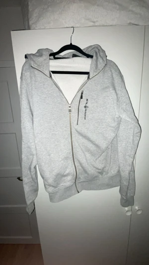 Grå hoodie från Sail Racing - Säljer en ljusgrå hoodie från Sail Racing med dragkedja och huva. Tröjan har en snygg logga på bröstet och en praktisk ficka med dragkedja. Perfekt för chill dagar och har en skön passform med långa ärmar.