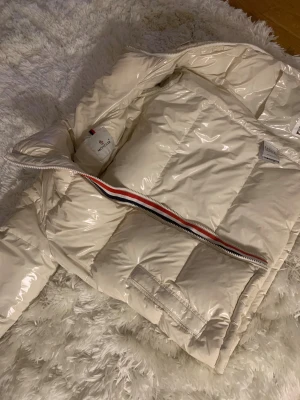 Beige Moncler pufferjacka med röd/vit/blå dragkedja - Säljer en beige pufferjacka från Moncler med glansig finish och ikonisk röd, vit och blå rand längs dragkedjan. Jackan har hög krage, fickor med lock och Moncler-logga på bröstet. Perfekt för dig som vill ha en clean och lyxig vinterstil.