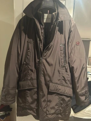 Brun Moncler dunjacka storlek L - Säljer en brun dunjacka från Moncler i storlek L. Jackan har hög krage, flera fickor med dragkedja och tryckknappar, samt justerbara ärmslut. Materialet är polyamid och fyllningen består av 80% dun. Perfekt för kalla dagar med sin långa och rymliga passform.