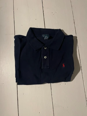 Mörkblå Polo Ralph Lauren piké - Snygg mörkblå pikétröja från Polo Ralph Lauren med klassisk krage och två knappar framtill. Tröjan har den ikoniska röda Polo-loggan broderad på bröstet och är tillverkad i 100% bomull för skön komfort.