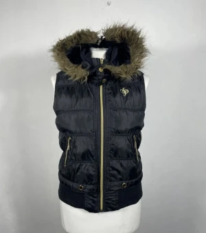 southpole väst - Svart pufferväst från southpole med guldfärgade dragkedjor och detaljer. Västen har en huva med fuskpäls och ribbade muddar nedtill. Perfekt för att addera lite edge till din outfit under kyliga dagar.