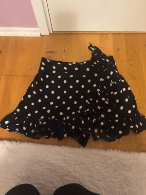 Svart prickig omlottkjol med volang - Supersöt svart kortkjol med vita prickar och volangkant. Kjolen har omlottmodell med knyt i midjan som ger en snygg och feminin look. Perfekt för dig som gillar lekfulla mönster och trendiga detaljer. finns även shorts under