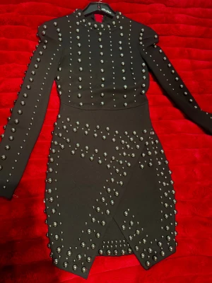 Svart kort klänning med nitar - Svart kort klänning med lång ärm och coola silverfärgade nitar över hela plagget. Klänningen har en omlott-design nedtill och markerade axlar som ger en edgy vibe. Perfekt för dig som vill sticka ut med en unik och trendig look.