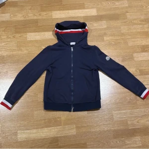 Marinblå Moncler hoodie med dragkedja - Snygg marinblå hoodie från Moncler med hel dragkedja och huva. Röd och vit rand på huvan och ärmsluten ger en sportig vibe. Tröjan har Moncler-logga på vänster ärm och två fickor framtill. Perfekt för dig som gillar stilrena märkesplagg. Kan till och med passa XS