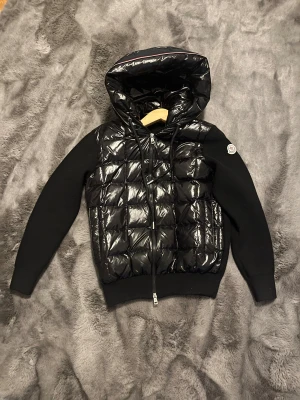 Svart quiltad pufferjacka från Moncler - Svart pufferjacka från Moncler med glansig quiltad front och huva, stickade ärmar och rygg. Jackan har dragkedja framtill, snörning vid huvan och Moncler-logga på vänster ärm. Perfekt för dig som gillar stilren och sportig look.