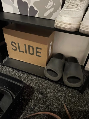 Adidas Yeezy Slide i mörkgrå - Säljer ett par Adidas Yeezy Slide i mörkgrå färg. Slidesen har en chunky och minimalistisk design med öppen tå och platt sula. Tillverkade i syntetmaterial för en bekväm och modern look. Perfekta för chill dagar eller när du vill ha något enkelt men trendigt på fötterna.