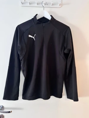 Svart långärmad träningströja Puma - Svart långärmad träningströja från Puma med vit logga på bröstet. Tröjan har en ståkrage och half zip-dragkedja framtill. Perfekt för sport eller chill, med en clean och sportig look. Materialet känns lätt och smidigt mot kroppen.
