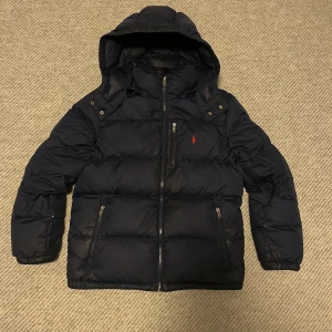 Mörkblå pufferjacka från Polo Ralph Lauren - Snygg mörkblå pufferjacka från Polo Ralph Lauren med röd broderad logga på bröstet. Jackan har huva, flera dragkedjeförsedda fickor och RL-detaljer på dragkedjorna. Perfekt för kalla dagar med sin vadderade design och klassiska look.
