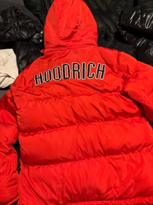Röd pufferjacka från Hoodrich - Säljer en fet röd pufferjacka från Hoodrich i storlek M. Jackan har huva, dragkedja framtill och stora fickor. Hoodrich-logga på bröstet och stor text på ryggen. 