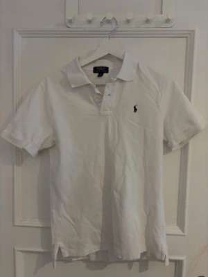 Vit pikétröja från Polo Ralph Lauren - Vit pikétröja från Polo Ralph Lauren med klassisk krage och korta ärmar. Tröjan har den ikoniska broderade loggan på bröstet och är tillverkad i 100% bomull för en skön och luftig känsla. Perfekt för en clean och stilren look. 