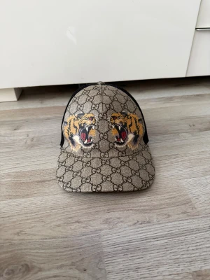 Gucci beige keps med tigertryck - Snygg keps från Gucci i beige med klassiskt GG-mönster och två coola tigerhuvuden framtill. Baksidan är i svart mesh med justerbart band i grönt och rött. Märkt med Gucci på insidan. Perfekt för dig som vill sticka ut med lyxig streetstyle. Endast använd 7 gånger!! ÄKTA!!!