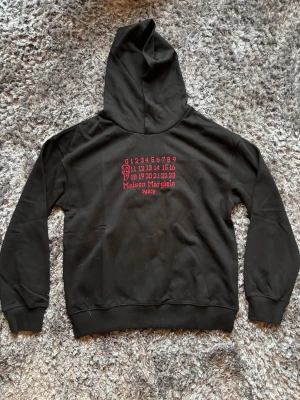 Maison Margiela svart hoodie med tryck - Svart hoodie från Maison Margiela med röd broderad logga och siffror på ryggen. Klassisk huva, ribbade muddar och de ikoniska fyra vita stygn i nacken. Mjuk bomullskvalitet som känns skön mot huden. Perfekt för dig som gillar streetwear med lyxig vibe.