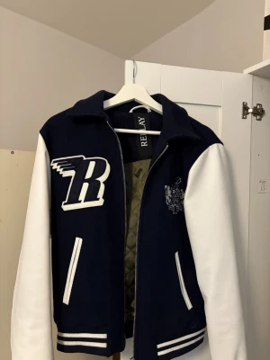 Blåvit bomberjacka från Replay - Snygg blå bomberjacka med vita ärmar och detaljer från Replay. Jackan har ett stort R-tryck på bröstet, broderad emblem, randiga muddar och dragkedja framtill. Insidan är quiltad i olivgrönt. Perfekt för dig som gillar college-stil. Ny pris 3200kr jag vill endast ha 1000kr