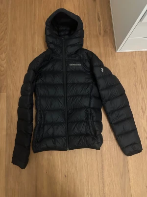 Svart dunjacka Peak Performance S - Svart dunjacka från Peak Performance i storlek S. Jackan har huva, quiltad design och dragkedja framtill. Perfekt för kalla dagar med sin puffiga look och stilrena svarta färg. Klassisk logga på bröstet och ärmen.