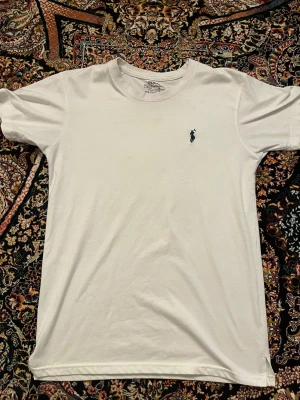 Vit t-shirt från Polo Ralph Lauren - Vit t-shirt från Polo Ralph Lauren i regular fit. Klassisk design med korta ärmar och rund halsringning. Liten mörkblå broderad logga på bröstet. Tillverkad i mjuk bomull som känns skön mot huden.