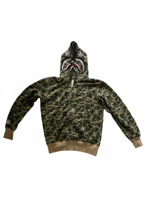BAPE Shark Camo Hoodie - Säljer en ikonisk BAPE Shark hoodie med grönt camomönster, hel dragkedja och broderad hajdesign på huvan. Huvan har även färgglada bokstäver och tigerpatch på sidorna. Klassisk streetwear med ribbade muddar och detaljer i bomull.
