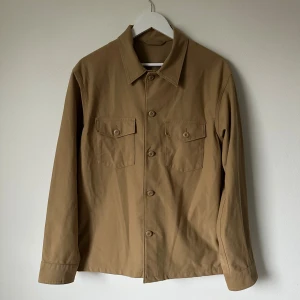 Beige overshirt från Uniqlo, M - Stilren beige overshirt från Uniqlo med klassisk krage och två bröstfickor med knappar. Jackan är tillverkad i en bomullsblandning med elastan för extra komfort. Perfekt för lager-på-lager och enkel att matcha med det mesta. 5N9