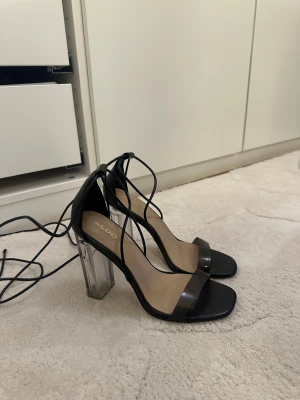 Svarta sandaler med transparent klack från Aldo - Snygga svarta sandaler från Aldo med transparent blockklack och tunna snörningar som går runt ankeln. Skorna har öppen tå och en bred rem i plast över foten. Perfekt för dig som gillar trendiga detaljer och vill sticka ut med en cool klack.