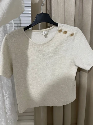 Krämvit topp med guldknappar - Säljer en krämvit topp i bouclé-liknande material med korta ärmar och rund hals. Toppen har tre snygga guldknappar på ena axeln som ger en cool detalj. Perfekt att styla med jeans eller kjol och kostymbyxor för en trendig look.