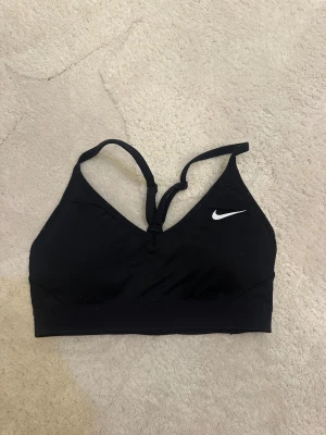 Svart sporttopp från Nike - Svart sporttopp från Nike med smala axelband och klassisk logga framtill. Toppen har en tight passform och är tillverkad i ett funktionsmaterial som andas, perfekt för träning. Snygg och enkel design med lätt stöd och elastisk nederkant.