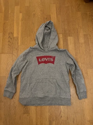 Grå Levi's hoodie med röd logga - Grå hoodie från Levi's i storlek M med klassisk röd logga på bröstet. Tröjan har huva, magficka och ribbade muddar vid ärmslut och nederkant. Perfekt för chill dagar och enkel att matcha med jeans eller joggers.