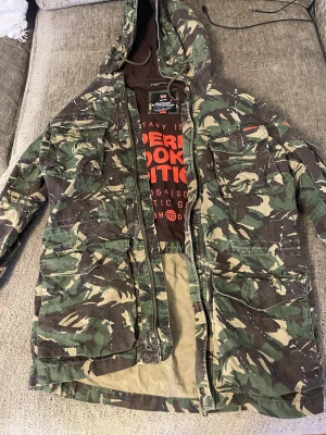 Camouflage parkajacka Superdry XL - Parkajacka från Superdry i militärinspirerat camouflage-mönster med gröna, bruna och beige toner. Jackan har huva med snörning, flera stora fickor med knappar och dragkedja framtill. Tuffa Superdry-loggor på bröst och ärm. Perfekt för dig som gillar streetstyle.
