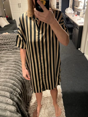 Randig skjortklänning från Black Label - Säljer en loose-fit skjortklänning från The Black Label Collection i storlek XS/S. Klänningen har vertikala svarta och beige ränder, korta ärmar och knappar hela vägen framtill. Perfekt för dig som gillar en chill och trendig look.