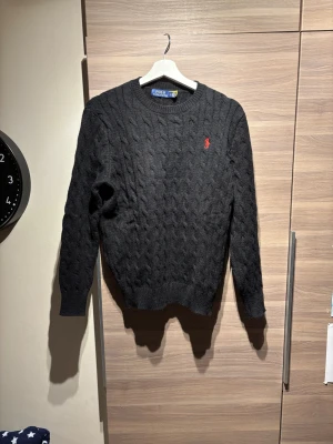 Svart stickad tröja Polo Ralph Lauren - Svart kabelstickad tröja från Polo Ralph Lauren med rund hals och lång ärm. Klassisk röd broderad logga på bröstet. Mjuk och varm känsla, perfekt för kyliga dagar. Ribbstickade muddar vid ärmslut och nederkant. Snygg att matcha med jeans eller chinos.