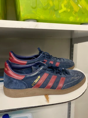 Adidas Spezial blå/röda sneakers mocka - Säljer ett par Adidas Spezial sneakers i mörkblå mocka med röda läderdetaljer och klassiska tre ränder på sidorna. Gummisula i brunt och guldfärgad Adidas-logga på sidan. Skorna har snörning och en retro vibe som passar perfekt till streetwear.