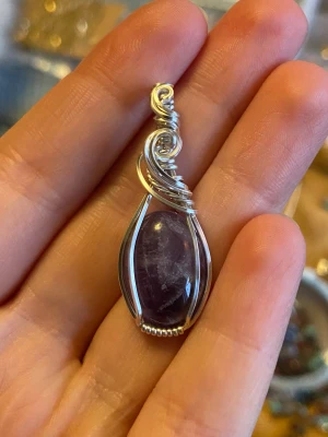 Handgjord ametist-hänge i silvertråd - Unikt hänge med en polerad, djup lila ametist infattad i konstnärligt virad silvertråd. Stenen är droppformad och omsluts av detaljerade spiraler och pärlformad kant. Perfekt för dig som gillar boho eller magisk stil.