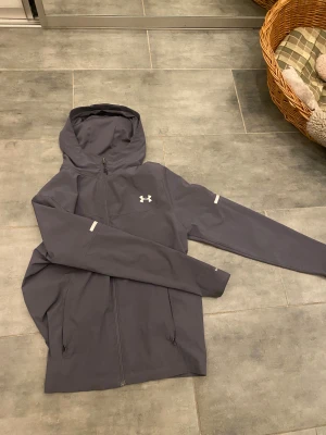 Grå vindjacka från Under Armour - Grå vindjacka från Under Armour med huva och dragkedja . Köpt ny och använd i 1 vecka 