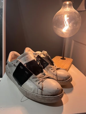 Valentino Garavani vita sneakers - Säljer ett par snygga vita sneakers från Valentino Garavani med bred svart detalj över sidan. Skorna har rund tå, platt sula och klassisk snörning. Tillverkade i skinn med coola detaljer och logga på plösen. Perfekt för dig som gillar exklusiv streetstyle.