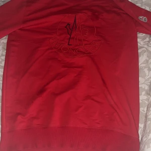 Röd sweatshirt från Moncler - Säljer en röd sweatshirt från Moncler med stort broderat logotyp på bröstet och patch på ärmen. Tröjan har rund hals och ribbade muddar vid ärmslut och nederkant. Perfekt för dig som gillar stilrena märkesplagg med tydlig branding.
