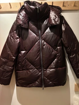 Mörkbrun pufferjacka Massimo Dutti - Snygg vintöd jacka från Massimo Dutti med hög krage och avtagbar huva som är i storleken XS. Jackan har quiltad design, dragkedja framtill och två sidofickor. Materialet är glansigt syntet och jackan har en loose passform. Jackan har fått en liten defekt på den vänstra ärmen men går att laga. Denna Massimo Dutti Jack är öerfekt nu inför vintern!                                       Orginal pris är 2300