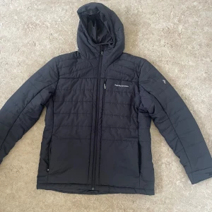 Svart pufferjacka Peak Performance XXL - Svart pufferjacka från Peak Performance i storlek XXL. Jackan har huva, quiltad design och flera praktiska fickor med dragkedja. Perfekt för kalla dagar och har en stilren look med broderad logga på bröstet och ärmen.