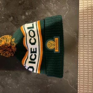 Grön och orange Jägermeister mössa - Snygg stickad mössa från Jägermeister i mörkgrönt med orange och vitt. Mössan har en stor tofs upptill och broderad patch med Jägermeister-logga framtill. Texten 'ICE COLD' syns på sidan. Perfekt för kalla dagar.