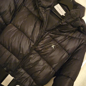 Svart pufferjacka från Calvin Klein - Svart pufferjacka från Calvin Klein Jeans med coolt bälte i midjan och CK-logga på bröstet. Jackan har dragkedja framtill och en snygg, quiltad design som ger extra volym. Perfekt för dig som vill ha en stilren och trendig look under kalla dagar.