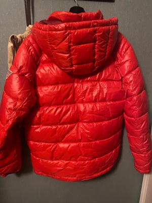 Röd pufferjacka från Guess - Säljer en röd pufferjacka från Guess med huva. Jackan har quiltad design och är perfekt för kalla dagar. Materialet är glansigt och känns riktigt skönt. Snygg logga på insidan och detaljer vid huvan.