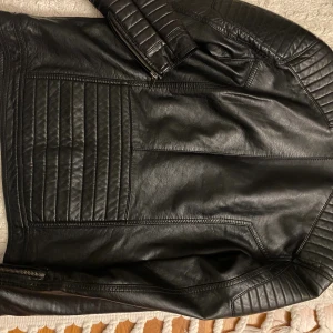 Svart skinnjacka från LCW Young XXS - Snygg svart skinnjacka från LCW Young i storlek XXS. Jackan har coola quiltade detaljer på axlar och armar, flera dragkedjor framtill och en ståkrage med knapp. Perfekt för dig som gillar en edgy stil och vill ha en jacka med attityd.