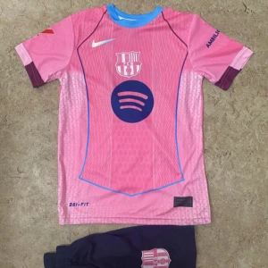 Barça Raphinha rosa fotbollströja Nike - Säljer en Barcelona fotbollströja i rosa med blå detaljer, korta ärmar och rund hals. Tröjan har tryck med Spotify-logga, FCB-emblem och Raphinha 11 på ryggen. Materialet är lätt och andas, Dri-Fit. Perfekt för dig som älskar Barça och vill sticka ut på planen.